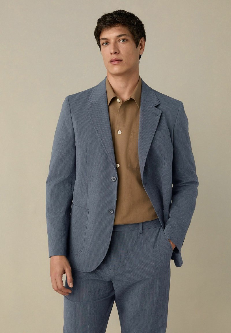 Giacca da sartoria azzurro chiaro con righe sottili, colletto a revers, chiusura con un solo bottone e tasca. Camicia marrone sotto con colletto e bottoni.
