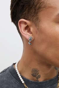 Silver earring featuring a skull with flames, hanging dice, worn on the lobe. The model also has a pearl necklace and a rose tattoo.