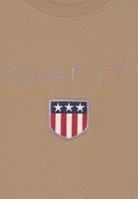Beżowa bluza z dużym logo "GANT" w jasnoszarym kolorze oraz wyszywanym herbem z trzema gwiazdami i czerwono-białymi paskami.