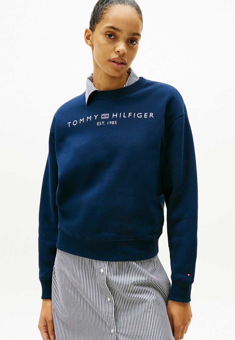 Felpa blu navy con polsini a costine, con logo bianco "TOMMY HILFIGER EST. 1985". Indossata sopra una camicia a righe con colletto. Texture liscia.