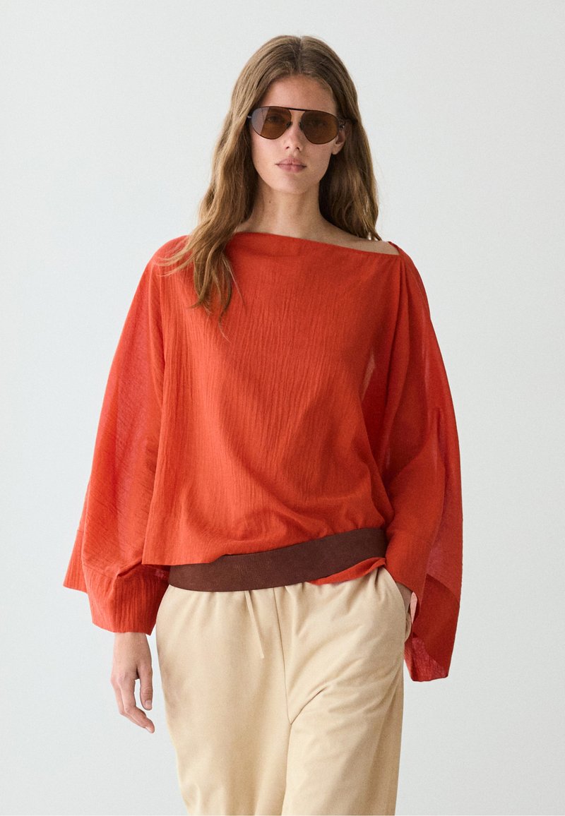 Vrouw met een bruine zonnebril, losse feloranje top met lange mouwen en één blote schouder, en beige broek tegen een effen lichte achtergrond.