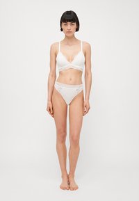 A fehér csipke bralette és a hozzá illő magas derekú bugyi puha kivitelezéssel, áttetsző csipke részletekkel és logós díszítéssel rendelkezik a derékpánton.