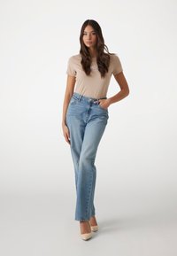 Camiseta ajustada beige con texto, jeans de tiro alto azul claros y tacones nude de punta. Modelo posando con una mano en la cadera, fondo neutral.
