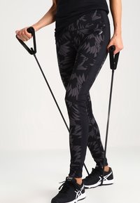 Henkilö, joka käyttää kuvioituja mustia legginssejä ja mustia urheilukenkiä vastusnauhan kanssa harjoitteluun.