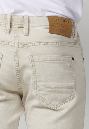 Beige denim jeans feature a straight cut, accented with a brown leather label at the waistband and subtle stitching on the back pockets.