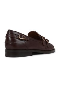 Brown leather slip-on shoe with a flat heel, featuring dual decorative gold-tone buckles and contrasting stitching on the side.