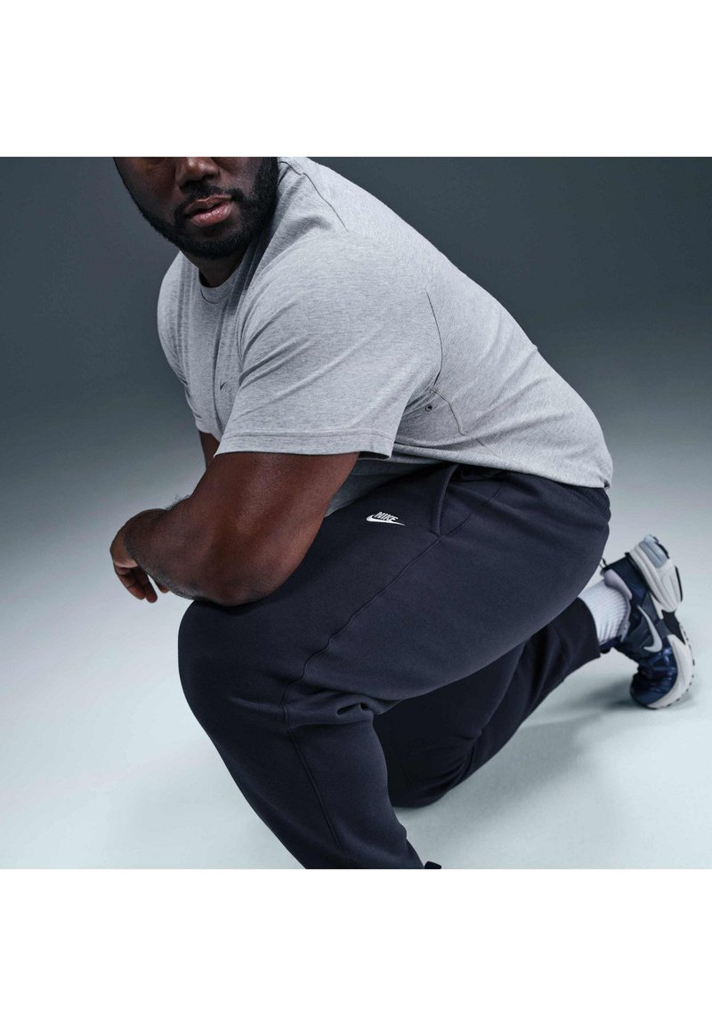Uomo con T-shirt grigia e pantaloni da tuta neri Nike in ginocchio su un ginocchio, di lato, con sfondo neutro e scarpe da ginnastica visibili.