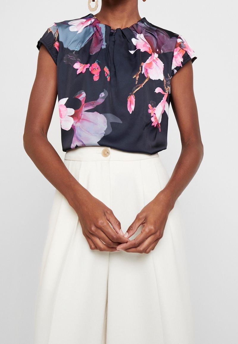 Blouse sans manches à motif floral foncé rentrée dans un pantalon blanc taille haute avec détail de boutons, portée par une personne en pose neutre avec des boucles d’oreilles pendantes.