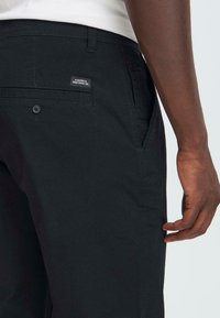 Vue partielle d'une personne portant un pantalon noir avec une poche arrière à bouton et une étiquette disant "Dédié à la perfection du denim".