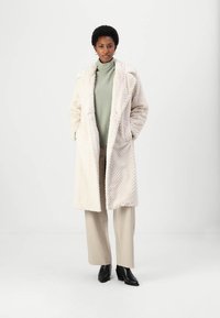 Langer weißer Kunstpelzmantel mit Zickzackmuster, Reverskragen und Seitentaschen, über einem hellgrünen Rollkragenpullover und beigefarbenen Hosen getragen.