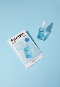 Torriden Dive In seerumi kirkkaassa lasipullossa tiputtimella, valkoisen pakkauksen vieressä, jossa on sinisiä yksityiskohtia, vaaleansinisellä taustalla.