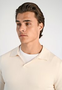 Krämfärgad stickad tröja med V-ringning och mjuk textur, med en kragdesigndetalj som ligger över en vit T-shirt.