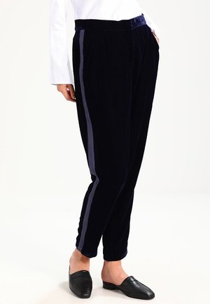 Person iført sorte fløjlsbukser med mørke sidelister og sorte slip-on sko, hænderne i lommerne, foran en ens hvid baggrund.