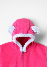 Růžová fleece bunda s předním zipem, kapucí s modrým chlupatým lemováním a dvěma kulatými modrými bambulemi na uších.