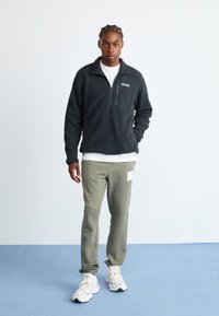 Man with braided hair wearing black fleece jacket, white T-shirt, olive green sweatpants, and white sneakers standing on blue floor against white wall.