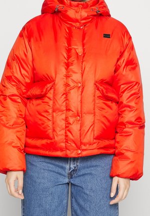 Doudoune rouge avec capuche, boutons-pression à l'avant, grandes poches avant, portée avec un jean bleu.