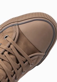 Zapatilla de cuero marrón con puntera de goma, cordones texturizados y suela estriada. Presenta una superficie lisa y detalles de costura reforzada.