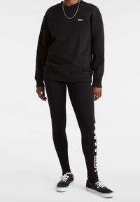 Person som bär en svart Vans sweatshirt, svarta leggings med vit Vans-logotyp på benet, svarta sneakers med vita snörning på vit bakgrund.