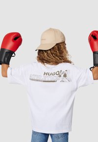 Camiseta blanca con estampado gráfico "HUGO ATHLETICS" en la parte posterior, gorra beige, guantes de boxeo negros y rojos, y pelo rizado visible.