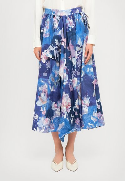 Femme portant une jupe midi plissée bleue à fleurs associée à des chaussures blanches à bouts pointus et un chemisier blanc à manches longues.