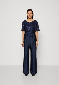 Mørkeblå jumpsuit med brede ben, med en dekorert kortermet topp og satengbånd i midjen. Glatt tekstur og skreddersydd passform.