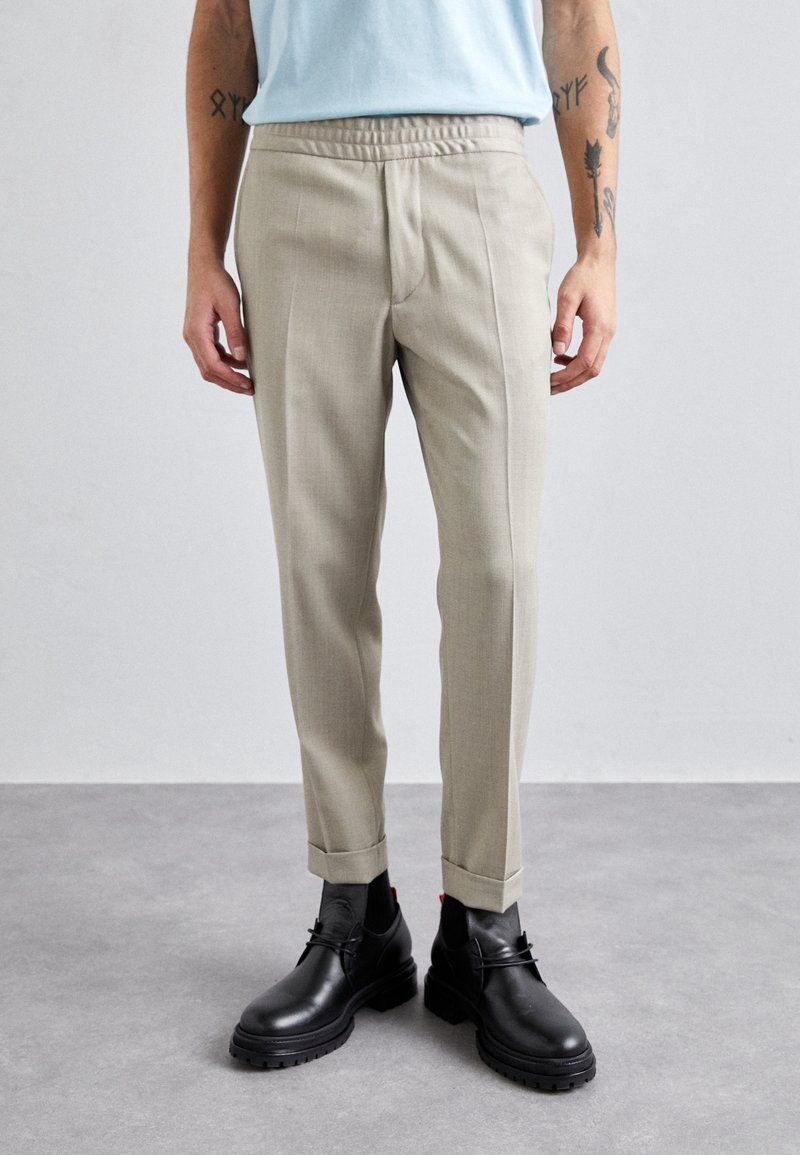 Homme portant un pantalon beige sur mesure avec des revers, des bottes en cuir noires à lacets et une chemise bleu clair, debout sur un sol gris.