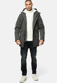 Mørkegrøn parka med hætte, to frontlommer og lynlåse. Bæres over en hvid striktrøje og sorte jeans, parret med lyse støvler.