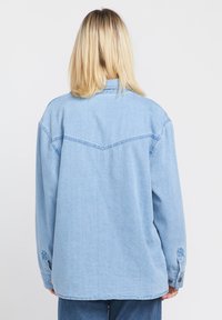 Lichtblauwe denimshirt met een rechte snit, spitse schouderdetails en knoopmanchetten. Zachte textuur met zichtbare stikselaccenten.