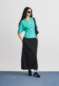 Γυναίκα με τιρκουάζ μπλουζάκι Puma, μαύρη μακριά φούστα, μαύρα αθλητικά παπούτσια Puma, γυαλιά ηλίου και τσάντα στον ώμο, στέκεται με τα χέρια στις τσέπες.