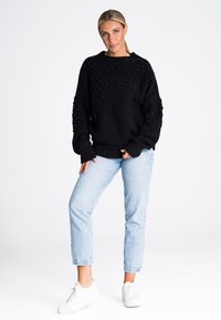 Czarny dziergany sweter z fakturowanym wzorem, okrągłym dekoltem i ściągaczami przy rękawach, w połączeniu z jasnoniebieskimi dżinsami i białymi sneakersami.