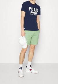 Navy blue cotton t-shirt with white "POLO 1967" print, paired with light green shorts and white sneakers with red and blue accents.