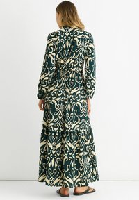 Long green dress with a beige abstract pattern, featuring long sleeves, a gathered waist, and a tiered skirt. Worn with black sandals.