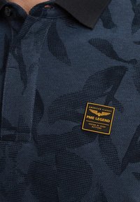 Polo shirt bleu marine avec un motif de feuilles texturé, col noir et une étiquette carrée PME Legend avec inscription dorée.