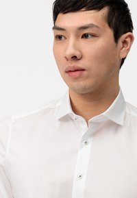 Chemise blanche à col italien, avec une texture en coton lisse et un léger éclat ; elle comporte deux boutons au niveau du col et un design ajusté.