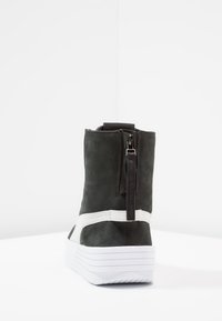 Svart mocka ankelsko med vita detaljer, bakre blixtlås och mönstrad gummisula. Har rundad tå och en något upphöjd plattform.