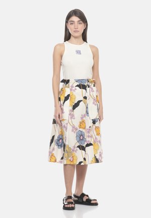 Donna in piedi che indossa un top bianco senza maniche con un piccolo logo e una gonna a fiori di media lunghezza con fiori gialli, blu e rosa, sandali neri.