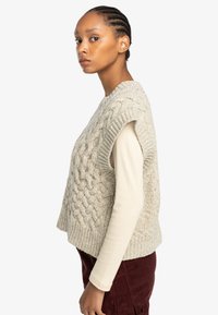 Light beige knitted vest with a textured cable pattern, short sleeves, worn over a cream long-sleeve top, showing a side profile.