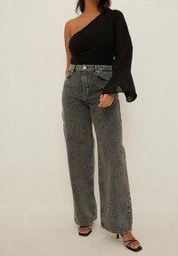 Svart topp med en axel och genomskinlig lång ärm, ihop med högmidjade, vida grå jeans och klackar med band i svart.