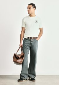 Lysfarvet tætsiddende t-shirt med logo, parret med løse grå jeans. Brun lædertaske og sorte slip-on sko med hvide accenter.