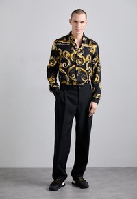 Camicia a maniche lunghe nera e oro con fantasia, pantaloni neri su misura, sneakers nere con accenti dorati, tessuto liscio, design aderente.