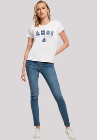 Weißes T-Shirt mit blauer Aufschrift "AHOI" und einem Anker-Graphic. Kombiniert mit engen blauen Jeans und weißen Sneakern. Lässiger Schnitt, kurze Ärmel.