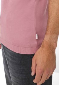Camicetta a maniche corte, aderente, di un rosa polveroso, realizzata in tessuto a maglia testurizzata, con orlo rigato e una piccola etichetta sul lato. Abbinata a pantaloni grigio scuro.