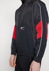 Svart nylonjacka med röda detaljer på ärmarna, vit kantning och framficka. Har hög krage och elastiska manschetter. Nike-logotypen syns.