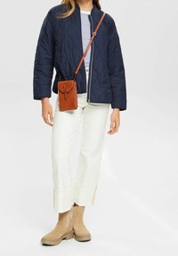Kobieta w granatowej pikowanej kurtce, w paski bluzce, białych szerokich spodniach z mankietami, beżowych botkach za kostkę i małej brązowej torebce na ramię.