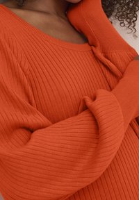 Maglione a coste con maniche lunghe di un vivace arancione. Presenta un ampio scollo e maniche morbide, evidenziando un tessuto morbido e texturizzato.
