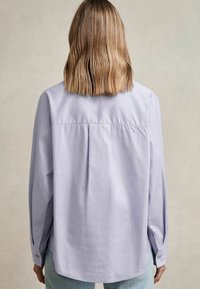 Light purple long-sleeve shirt with a pleated back, buttoned cuffs, and a relaxed fit. Fabric appears smooth, with a slight sheen.