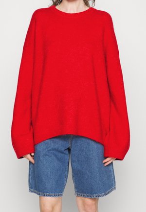 Persoon draagt een oversized rode trui met lange mouwen en blauwe spijkershorts tot halverwege de dijen, tegen een effen lichte achtergrond.