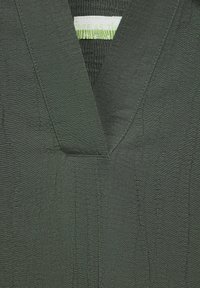 Tissu vert foncé avec une finition texturée, mettant en vedette un col en V et des coutures contrastées. L'étiquette indique "Libre comme le..." au niveau du col.