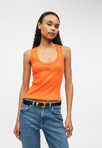 Narancssárga bordázott ujjatlan póló, mély dekoltárral, kék farmernadrággal és fekete övvel, amelynek arany csatja van.