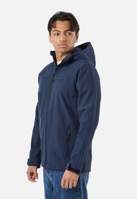 Jeune homme portant une veste à capuche bleu marine avec fermeture éclair à l'avant et poches zippées, debout devant un fond blanc uni.